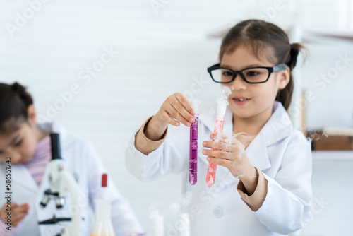 In chemistry classroom with many laboratory tools. Two little Asian girl in white lab coat help each other for experiment. A girl with glasses pick up orange and violet solution test tubes carefully.