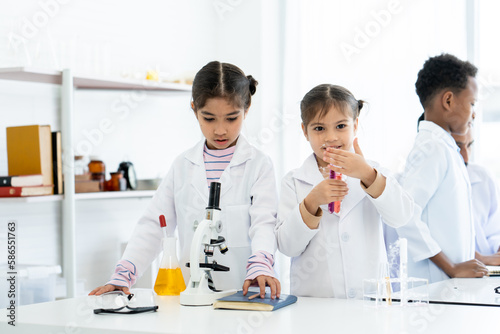 In chemistry classroom with many laboratory tools. Students and teacher do experiment together. Two little Asian girls help each other for preparing solution in test tubes and yellow chemical flask.