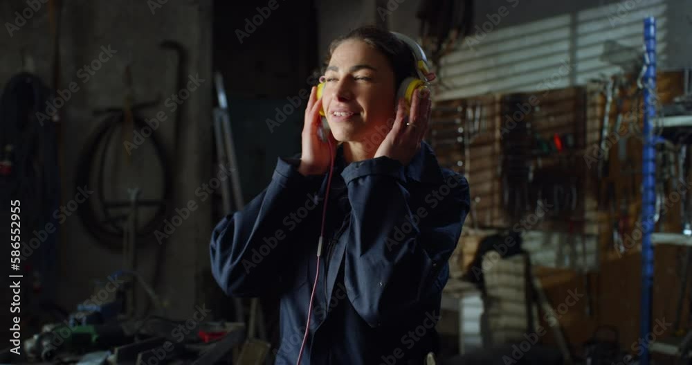 Portrait of Young Female Mechanic in Uniform Overalls Putting Music Headphones on and Listening to Happy Music. Professional Woman Working as a Welder Taking a Break in her Maintenance Workshop Portrait of Young Female Mechanic in Uniform Overalls Putting Music Headphones on and Listening to Happy Music. Professional Woman Working as a Welder Taking a Break in her Maintenance Workshop