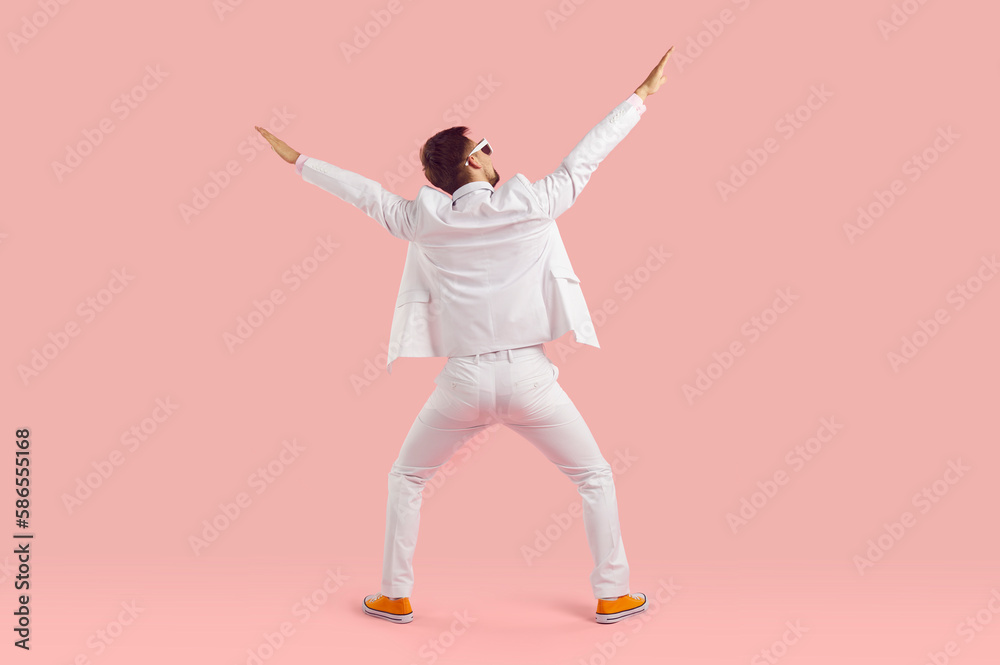 Fototapeta premium Funny inspired man in white suit performs energetic fashionable dance stands with back to camera stretching arms up and hangs out having carefree time stands on studio pink background