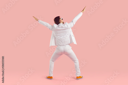 Funny inspired man in white suit performs energetic fashionable dance stands with back to camera stretching arms up and hangs out having carefree time stands on studio pink background