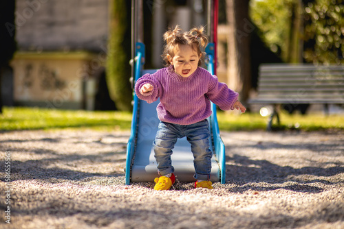 Sweet caucasian little toddler girl with two tails having fun smiling widely sliding down the yellow hill head first in the playground with violet sweeter sleeveless jacket jeans photo