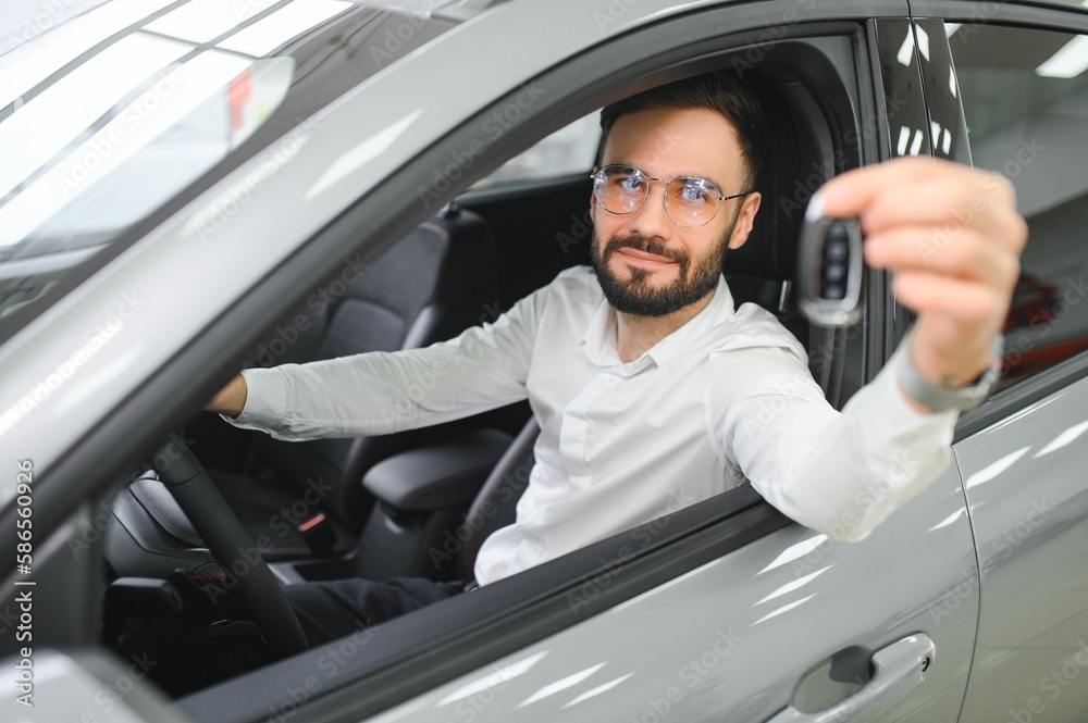 Handsome young businessman in classic white suit is smiling, looking at camera and showing car keys while sitting in car in a motor show. Handsome young businessman in classic white suit is smiling, looking at camera and showing car keys while sitting in car in a motor show.