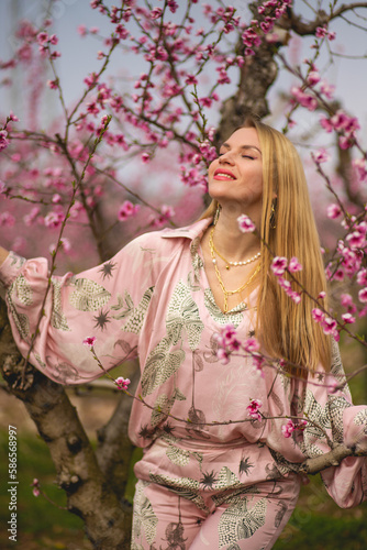 The portrait of young sensual blondie woman in pink silk costume smiling and enjoying the blossom peach garden 