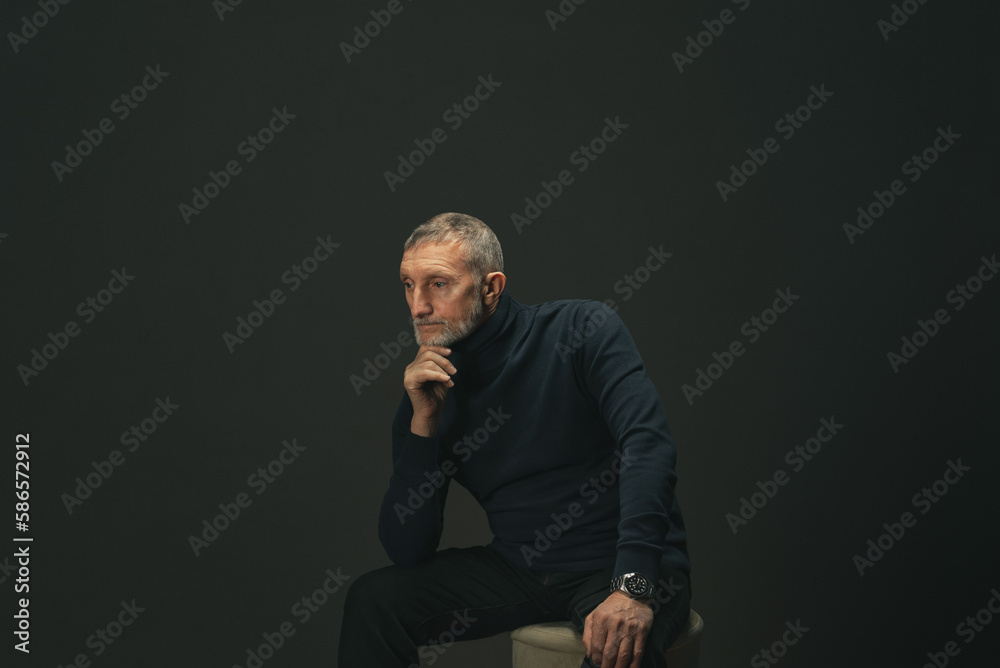an elderly man with a beard is sitting on a pouf in the studio on a dark background, he was thinking about something an elderly man with a beard is sitting on a pouf in the studio on a dark background, he was thinking about something