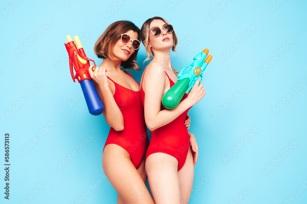 Two beautiful sexy smiling hipster women in red summer swimwear bathing suits. Trendy models having fun in studio. Hot female isolated n blue. Cheerful and happy. Playing toy water gun Two beautiful sexy smiling hipster women in red summer swimwear bathing suits. Trendy models having fun in studio. Hot female isolated n blue. Cheerful and happy. Playing toy water gun