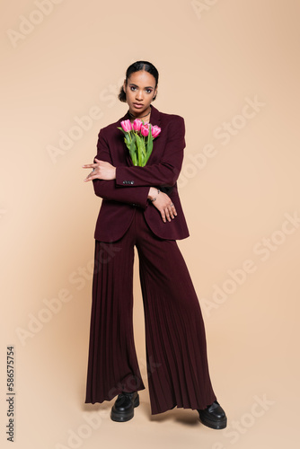 full length of well dressed african american woman in burgundy suit posing with pink tulips on beige.