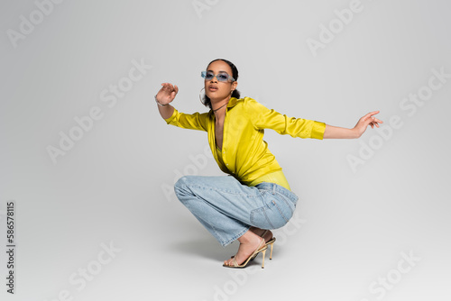 Photo full length of stylish african american woman in high heels and trendy outfit sitting on haunches on grey