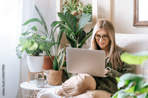 Freelance woman in glasses typing at laptop and working from home office. Happy girl sitting on couch in living room with plants in cozy urban jungle home. Distance learning online education and work.