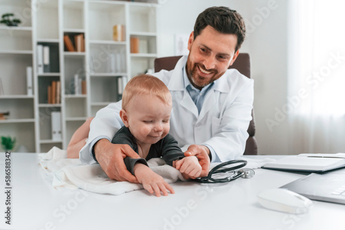 Toddler is lying down on the table. Doctor with little baby is working in the cabinet © standret