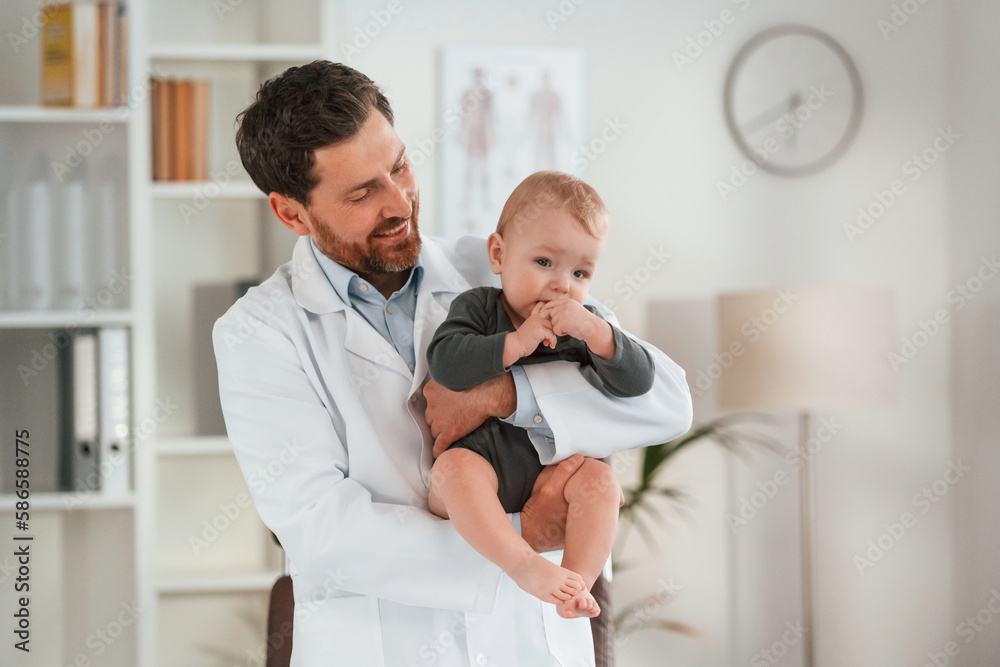 Fototapeta premium Standing and holding boy. Doctor with little baby is working in the cabinet