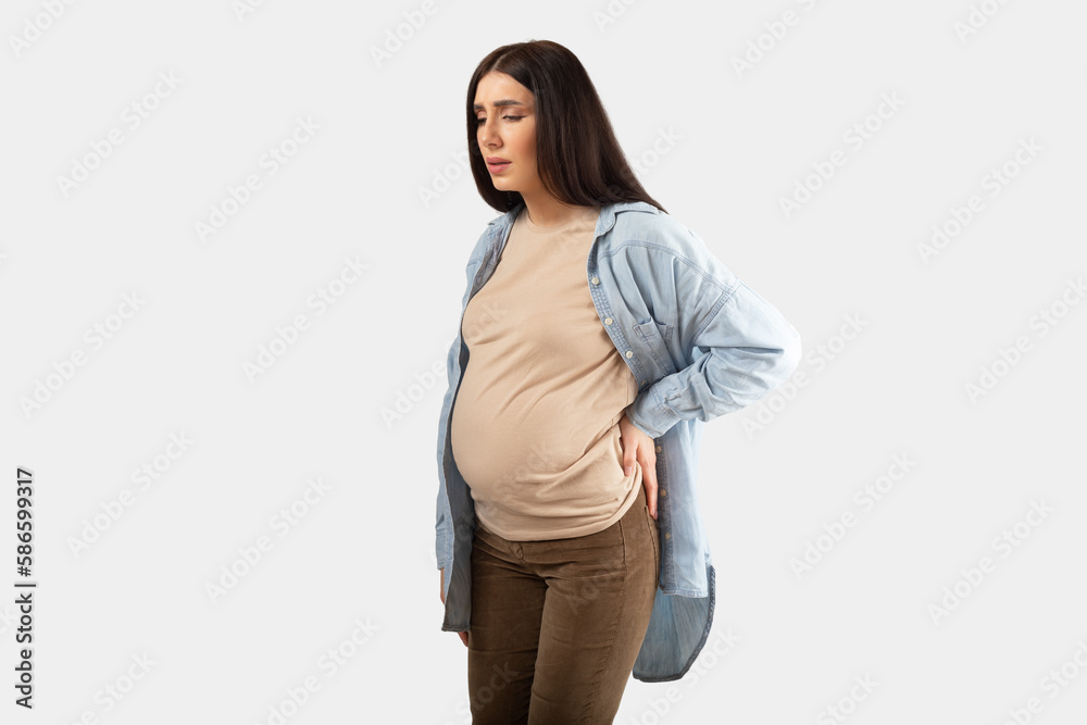 Pregnancy backache. Portrait of pregnant woman suffering lower back pain, feeling unwell, standing on white background Pregnancy backache. Portrait of pregnant woman suffering lower back pain, feeling unwell, standing on white background