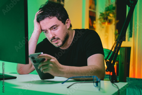 Young latin man sitting at his desk while looking at his cell phone worriedly. photo