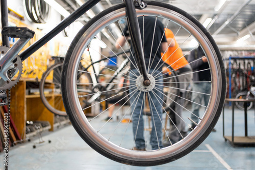 detail of a bicycle wheel hanging in a workshop, through the spokes we can see two unrecognizable men fixing another bike.
