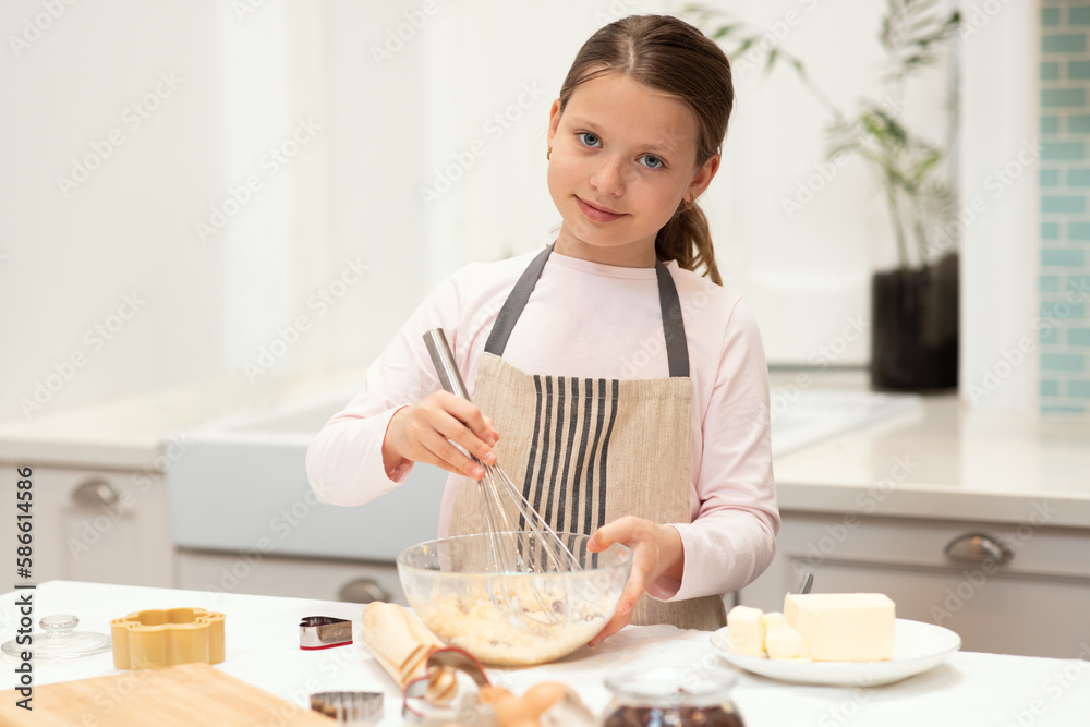 Cheerful caucasian little girl in apron make cookie dough with whisk in kitchen interior, copy space