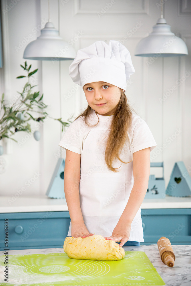 little cute smiling girl wearing a white chef's hat and uniform with tongue kneading the dough for cake in the blue and white kitchen with easter decoration little cute smiling girl wearing a white chef's hat and uniform with tongue kneading the dough for cake in the blue and white kitchen with easter decoration