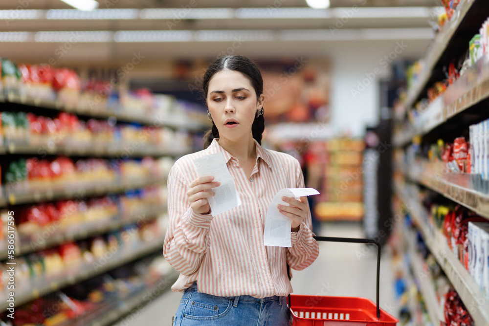 Portrait of young amazement Caucasian woman by high prices in supermarket. Shocked millennial holds receipts of purchase. In background, rows of shelves with products. Concept of inflation
