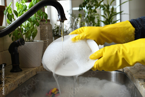 Washing rinsing dishes in kitchen sink by hand wearing yellow rubber gloves.