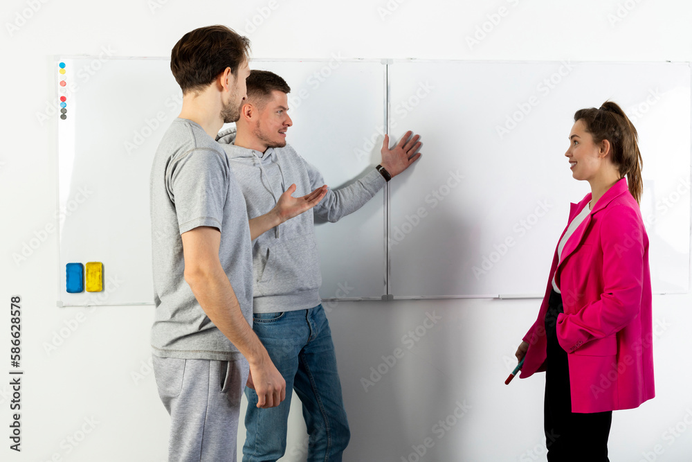 Fototapeta premium A team of three people standing against a whiteboard, communicating and discussing business. IT team in the office, two men and a woman. Young serious entrepreneurs discussing business.