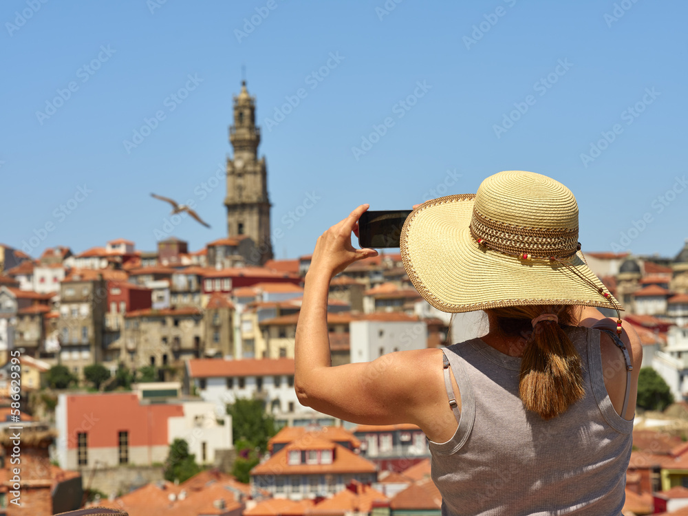 Woman wearing a straw hat to protect herself from the sun photographing the panorama of the historic city of Porto, Portugal. In the background the Torre de los Clérigos