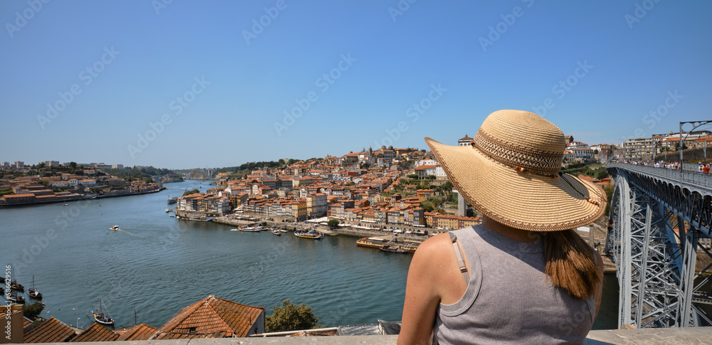 Woman wearing a straw hat to protect herself from the sun looking at the panorama with the Douro river and the historic city of Porto, Portugal. Dom Luis 1 iron bridge