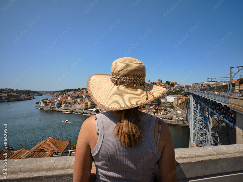 Woman wearing a straw hat to protect herself from the sun looking at the panorama with the Douro river and the historic city of Porto, Portugal. Dom Luis 1 iron bridge