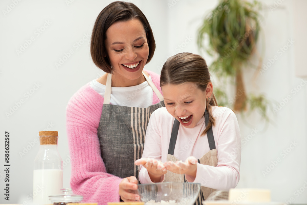 Satisfied caucasian young mom and small daughter in aprons make dough for cookies with flour, have fun Satisfied caucasian young mom and small daughter in aprons make dough for cookies with flour, have fun