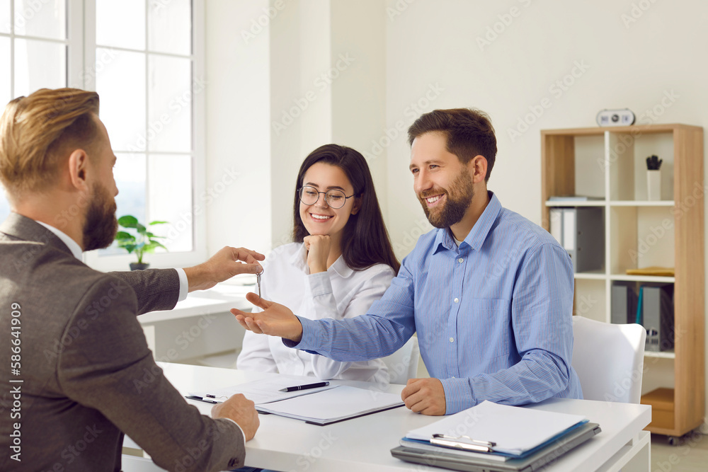 Realtor giving key to new apartment to young couple. Real estate agent in classic suit giving house keys to man and woman and signing agreement in office. Happy couple bought, rented new house Realtor giving key to new apartment to young couple. Real estate agent in classic suit giving house keys to man and woman and signing agreement in office. Happy couple bought, rented new house