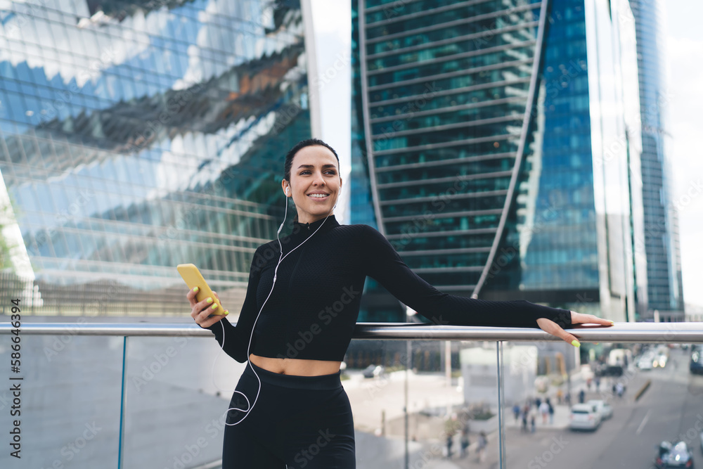 Cheerful standing sportswoman listening to music in earphones