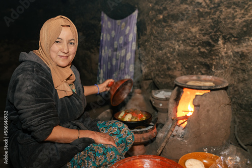 Moroccan berber woman in typical old kitchen with wood oven looking at camera and showing food in tajin