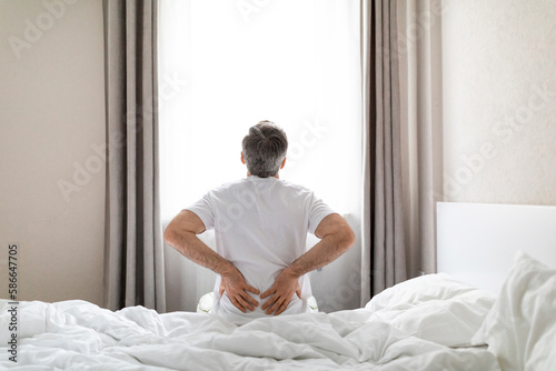 Back view of grey-haired man sittting on bed, touching back