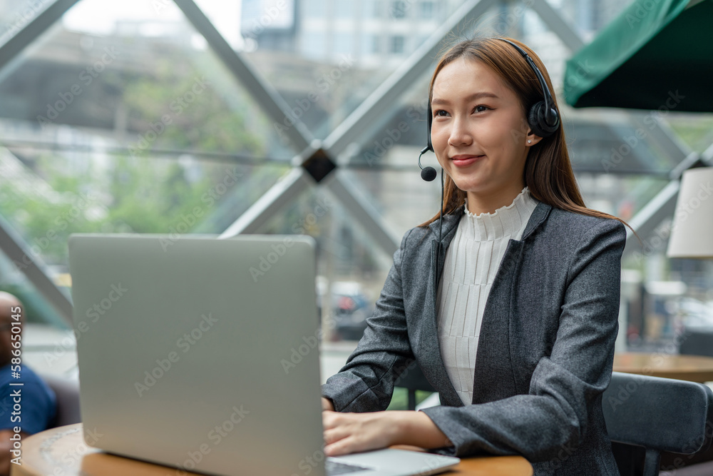 Young asian businesswoman working on her computer laptop and headset microphone in a cafe. Customer service executive working out of home. Hybrid workplace. Young asian businesswoman working on her computer laptop and headset microphone in a cafe. Customer service executive working out of home. Hybrid workplace.