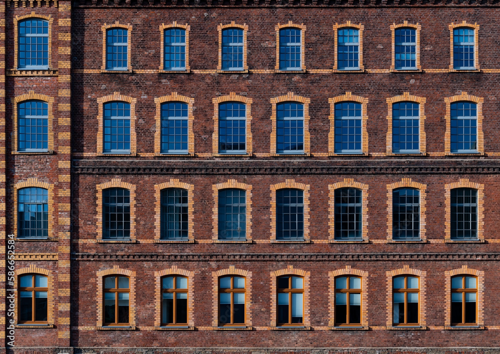 Historic brick facade of an ancient factory building in Iserlohn Sauerland Germany. Regular grid of big windows in four floors. Red and yellow brick wall with ornaments and glass reflecting blue sky.