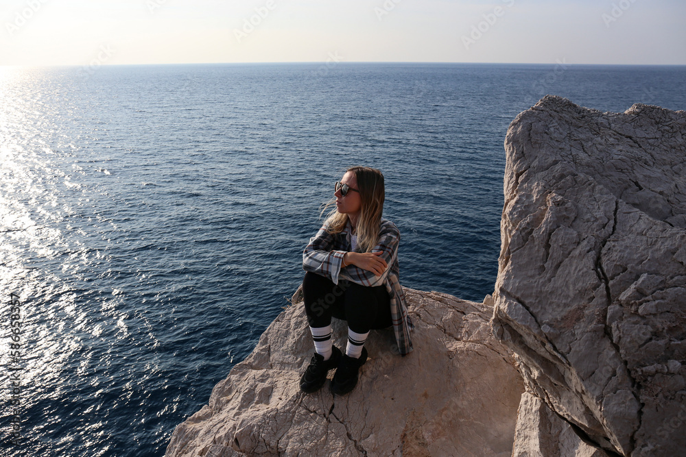 Young woman wearing sunglasses sitting on the edge of a cliff and enjoying the view of rocky coastline of atlantic ocean. Female connecting with nature. Copy space, background.