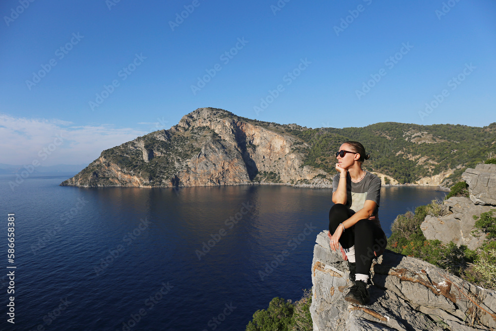 Young woman wearing sunglasses sitting on the edge of a cliff and enjoying the view of rocky coastline of atlantic ocean. Female connecting with nature. Copy space, background.
