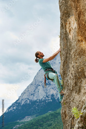 Active young man climbing on height vertical rock with rope. Gorgeous confident male climber looking up and overcoming a difficult route on cliff