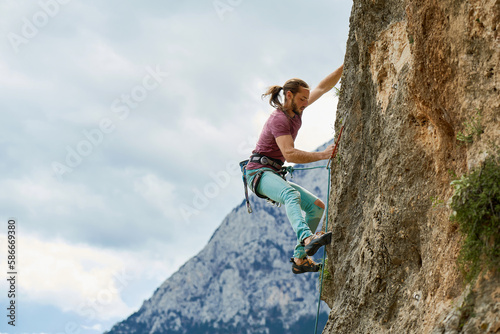 Side view strong concentrated man climbing on vertical rock with rope, lead climbing. Working on challenging route on cliff, beautiful mountains view on background