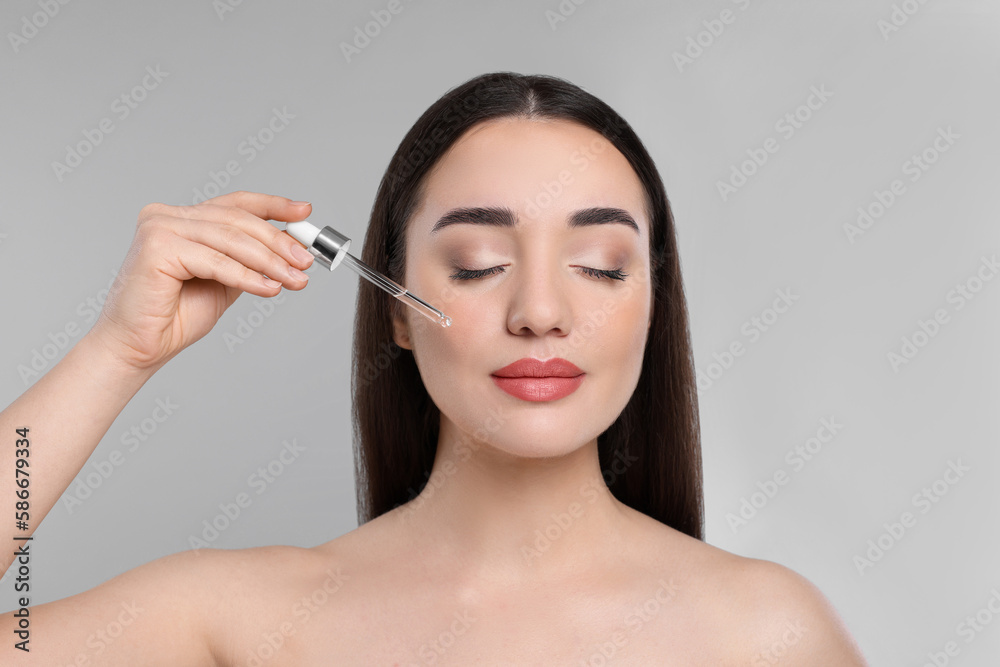 Young woman applying essential oil onto face on light grey background