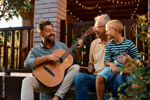 Happy man plays acoustic guitar to his son and senior father in backyard.