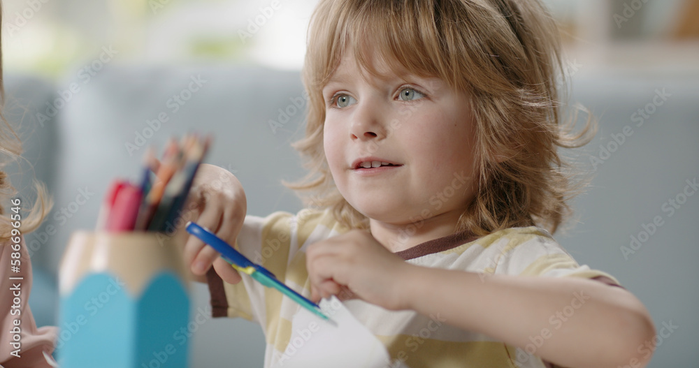 Funny little kid playing while crafting something. Cute boy cutting paper with scissors for children and happily smiling - happy childhood concept Funny little kid playing while crafting something. Cute boy cutting paper with scissors for children and happily smiling - happy childhood concept