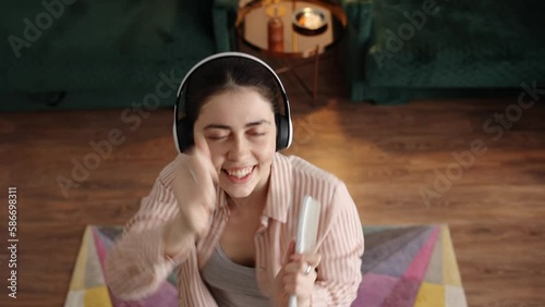 Top view of happy young Caucasian woman wearing headphones dancing and singing with hairbrush and looking at camera. Zoom in shot. Home weekend party