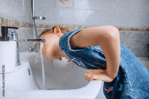 A little girl in a denim dress with a print leaned on a white ceramic sink to drink water from the tap in the bathroom.