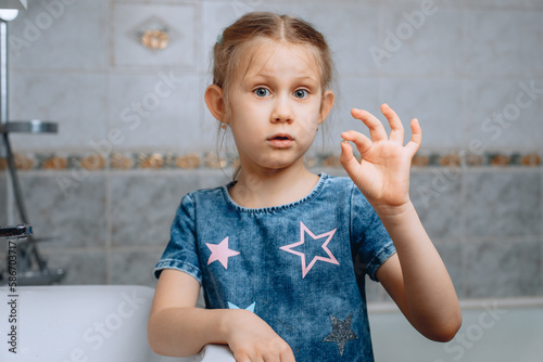 A surprised little preschool girl holds a tiny baby tooth in her hand while standing in the bathroom.