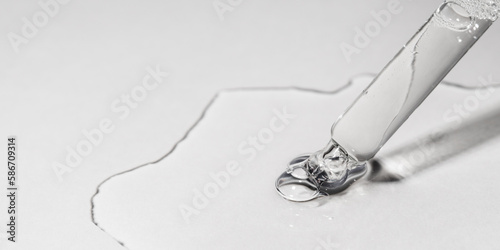 Drop of facial serum or oil with bubbles pouring from transparent pipette on grey background. Web banner for cosmetic product advertising. Liquid gel with retinol or squalane oil closeup. Front view.