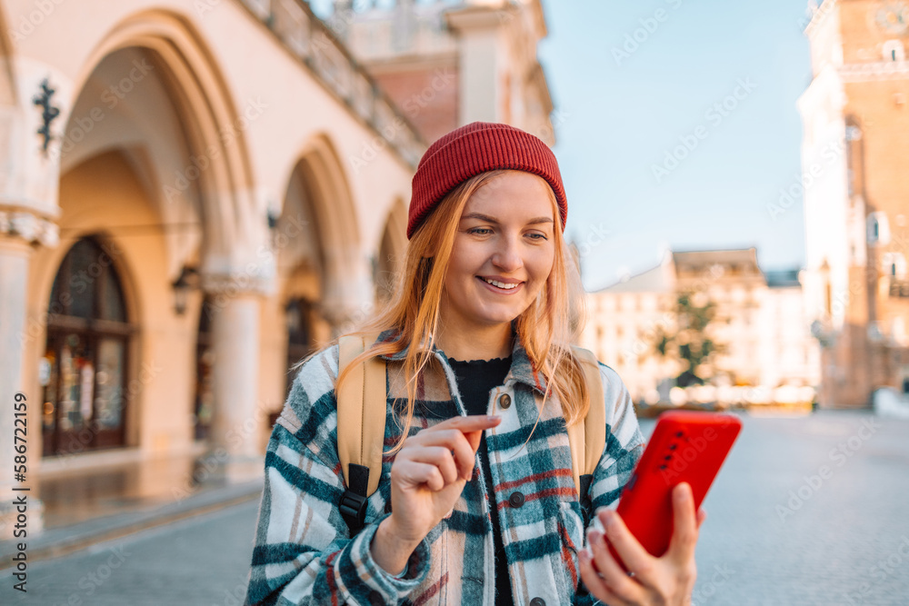Outdoor shot of pretty beautiful blonde haired young woman uses smartphone technology for writing web review browses website wears spectacles and raincoat carries bag poses. High quality photo