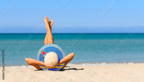 A slender girl on the beach in a straw hat in the colors of the Aruba flag. The concept of a perfect vacation in a resort in the Aruba. Focus on the hat.