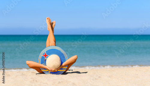 A slender girl on the beach in a straw hat in the colors of the Aruba flag. The concept of a perfect vacation in a resort in the Aruba. Focus on the hat. photo