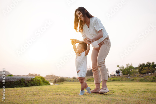 Baby taking first steps in park on meadow grass at sunset. Mother supports child to learn walking forward, baby growth and development concept.