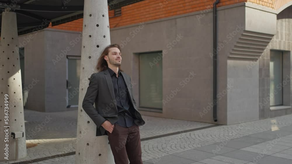 Handsome guy with beard and long hair is standing outdoor on office building background. He wears gray jacket, jeans. He looks to the side. man looking dreamy Handsome guy with beard and long hair is standing outdoor on office building background. He wears gray jacket, jeans. He looks to the side. man looking dreamy