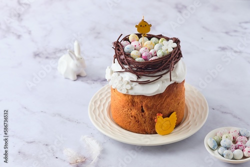 Easter cake covered with icing with a decor of a chocolate nest with eggs and a chicken topper on a white plate on a marble background. Easter table, treats, decor, traditions concept.